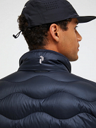 Back and side view of a person wearing a navy quilted jacket with a high collar and a black baseball cap against a light background.