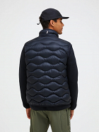 Back view of a man wearing a dark quilted puffer vest over a black fleece top, with a black cap and olive pants, on a white background.
