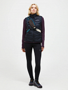 Full-body view of a woman wearing a black quilted zip vest, purple long-sleeve top, black leggings, sneakers, and a crossbody sling bag.