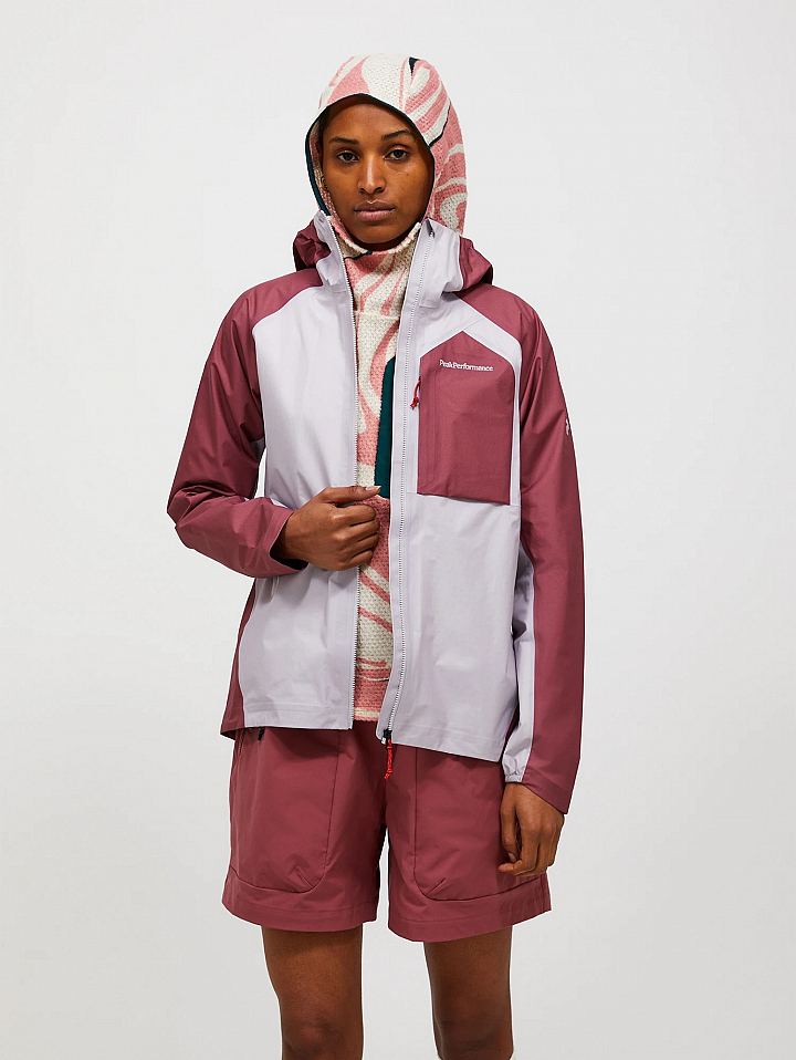 Model in a mauve and light gray hooded jacket, zipped partly open, with mauve shorts and a patterned hood against a white background.