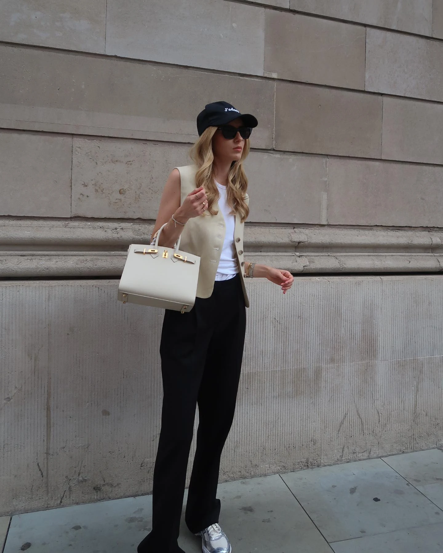 Valeria Kei wearing a cream vest, white top, black wide-leg trousers, cap, and sneakers on a London street
