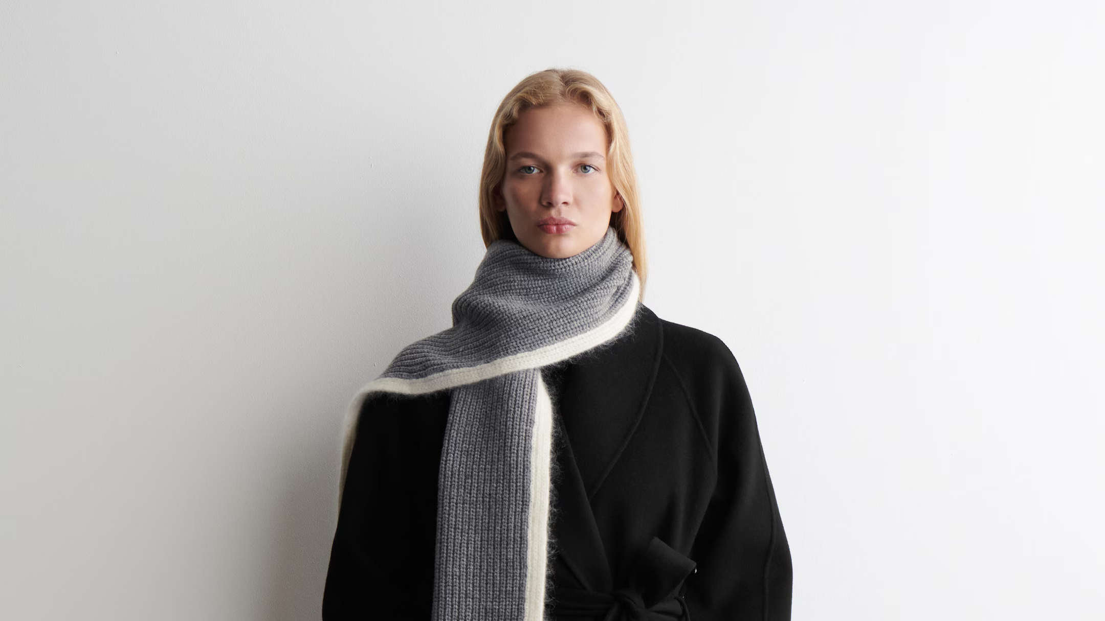 A model in a black coat wears a chunky grey scarf wrapped high around the neck with one long directional tail