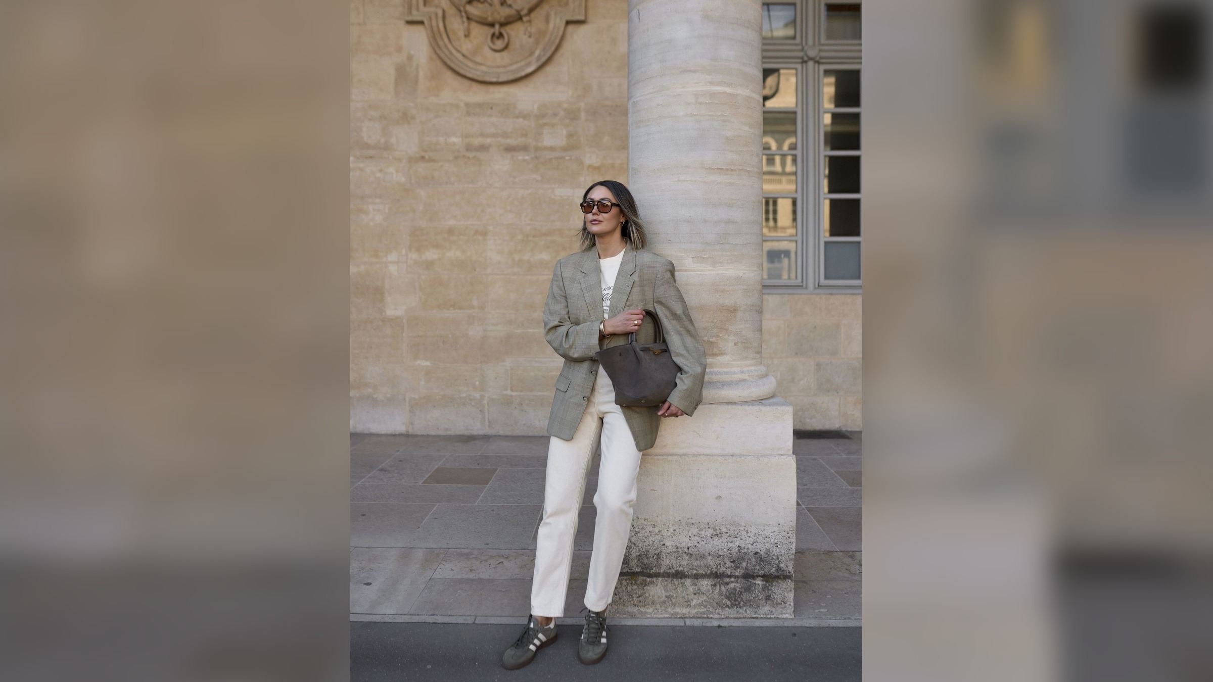 A woman in an oversized checked blazer, straight white trousers, defined sneakers, and a structured dark bag