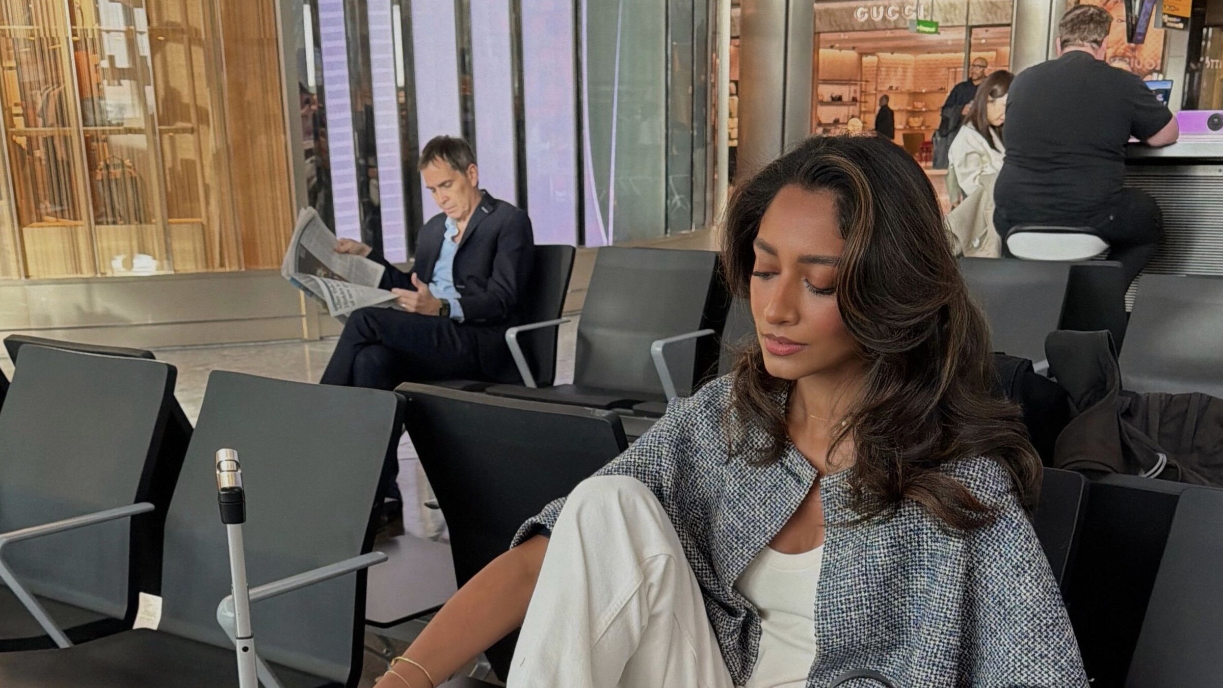 Traveler in white trousers and a textured gray jacket seated beside a suitcase at an airport